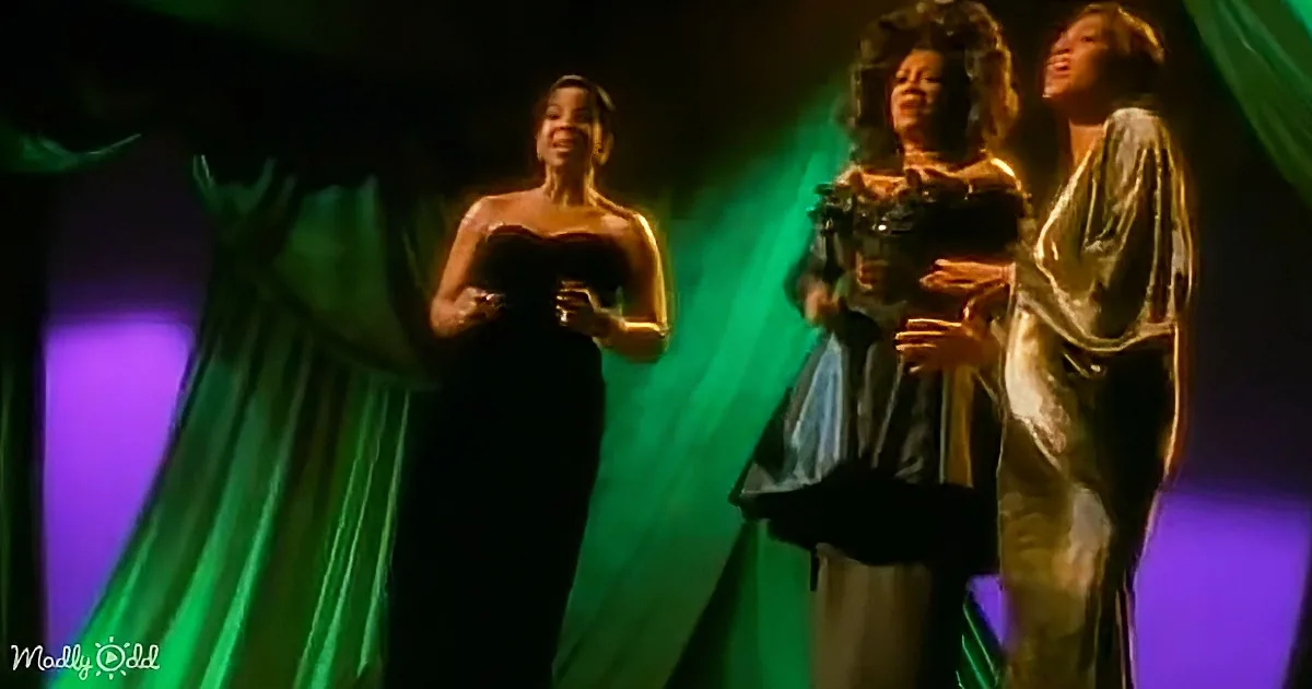Three women in elegant dresses stand on stage, one holding a bouquet, under green and purple lighting.