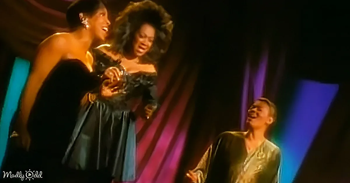 Two women in elegant dresses share a joyful moment on stage, while a man in a suit looks on with amusement.
