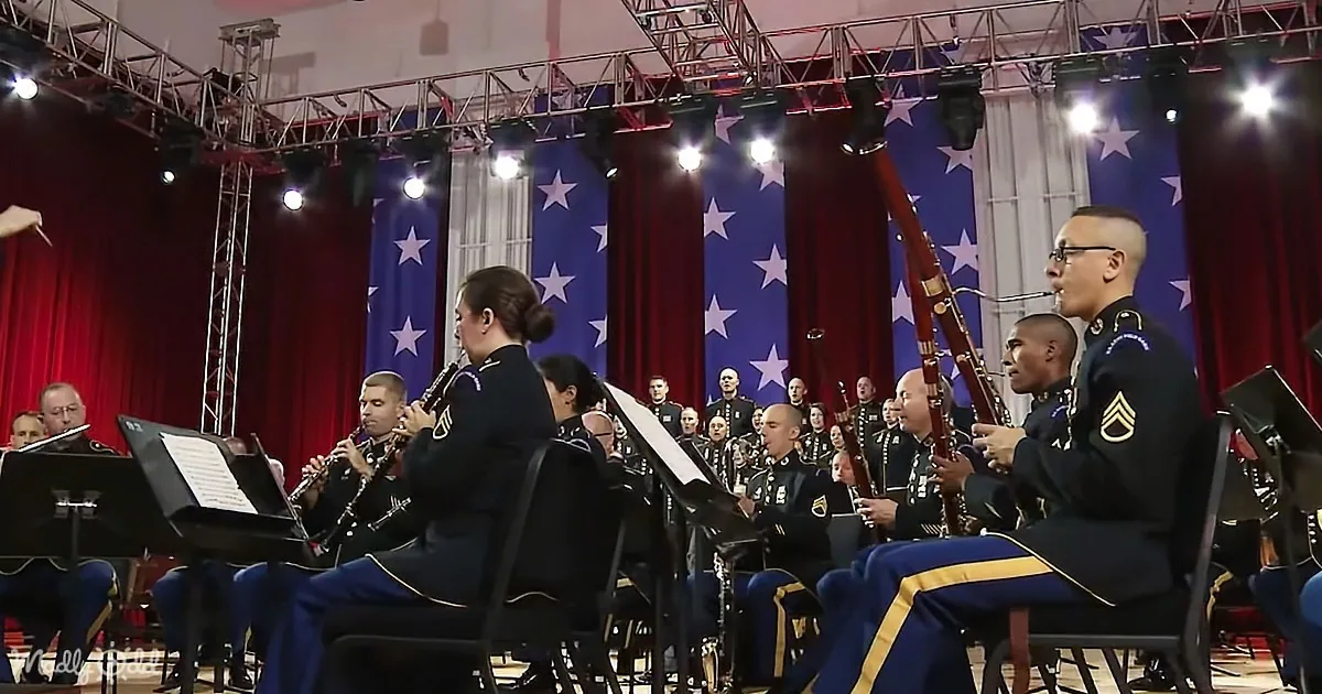 A military band in uniform plays instruments on stage with American flags in the background.
