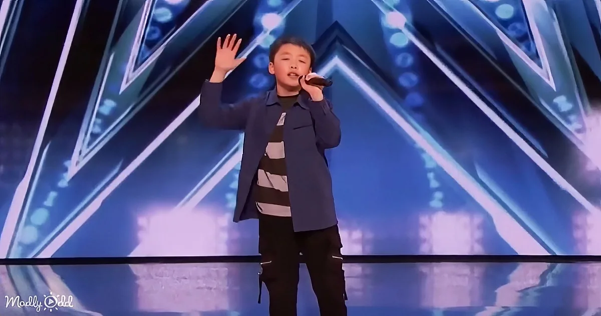 A young boy in a blue jacket and striped shirt sings on stage with a microphone, raising one hand.