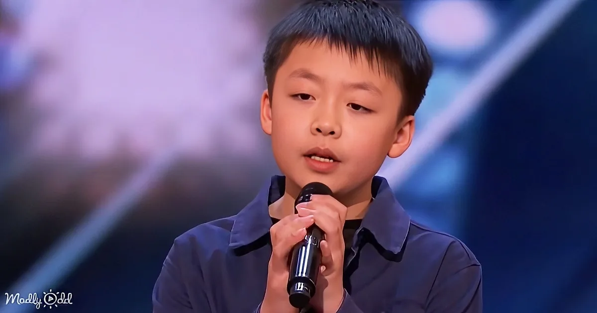 A young boy in a dark shirt holds a microphone, standing on a stage with a blurred background.