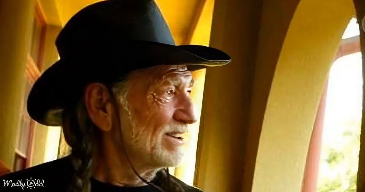 An older man with braided hair, wearing a black hat, smiles while standing in a warmly lit room with arched windows.