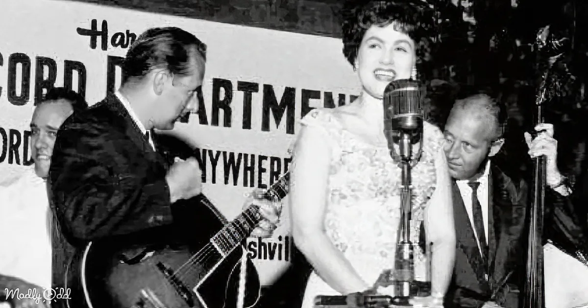 A woman sings into a microphone, accompanied by a guitarist, with a sign in the background.