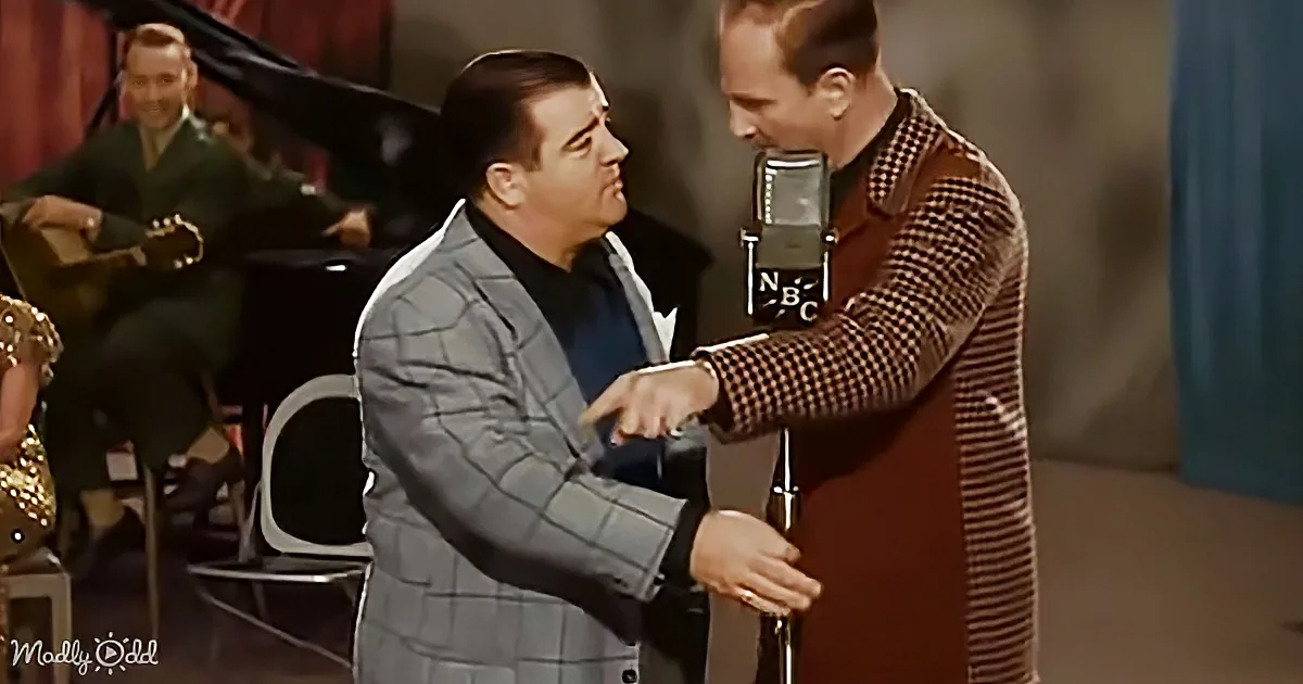 A man in a plaid suit shakes hands with another man in a brown checkered suit, both near a microphone.