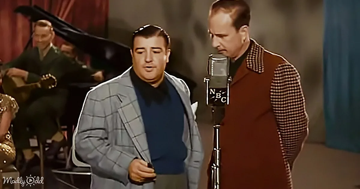 Two men in vintage attire sing into a microphone, one holding a cigarette, with a band in the background.