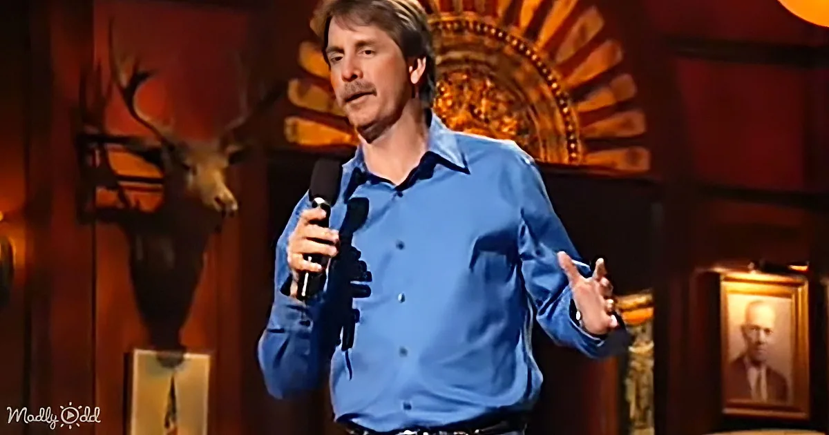 A man in a blue shirt holds a microphone, gesturing while speaking on stage with a deer head and ornate background.