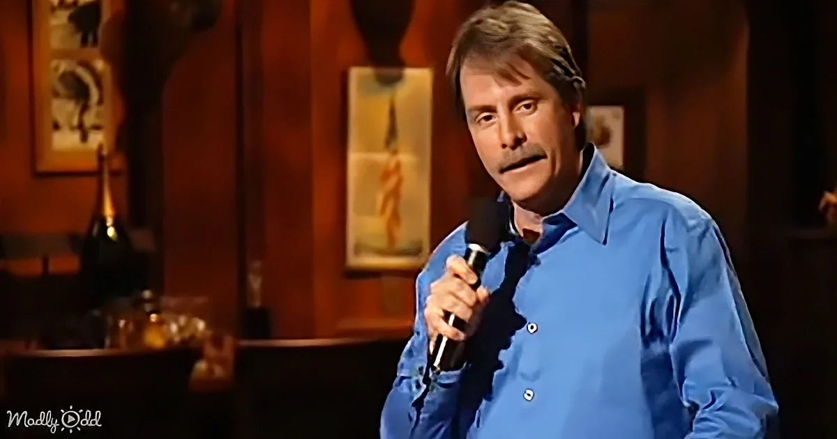 A man in a blue shirt holds a microphone, speaking on stage with a dark background and framed art.