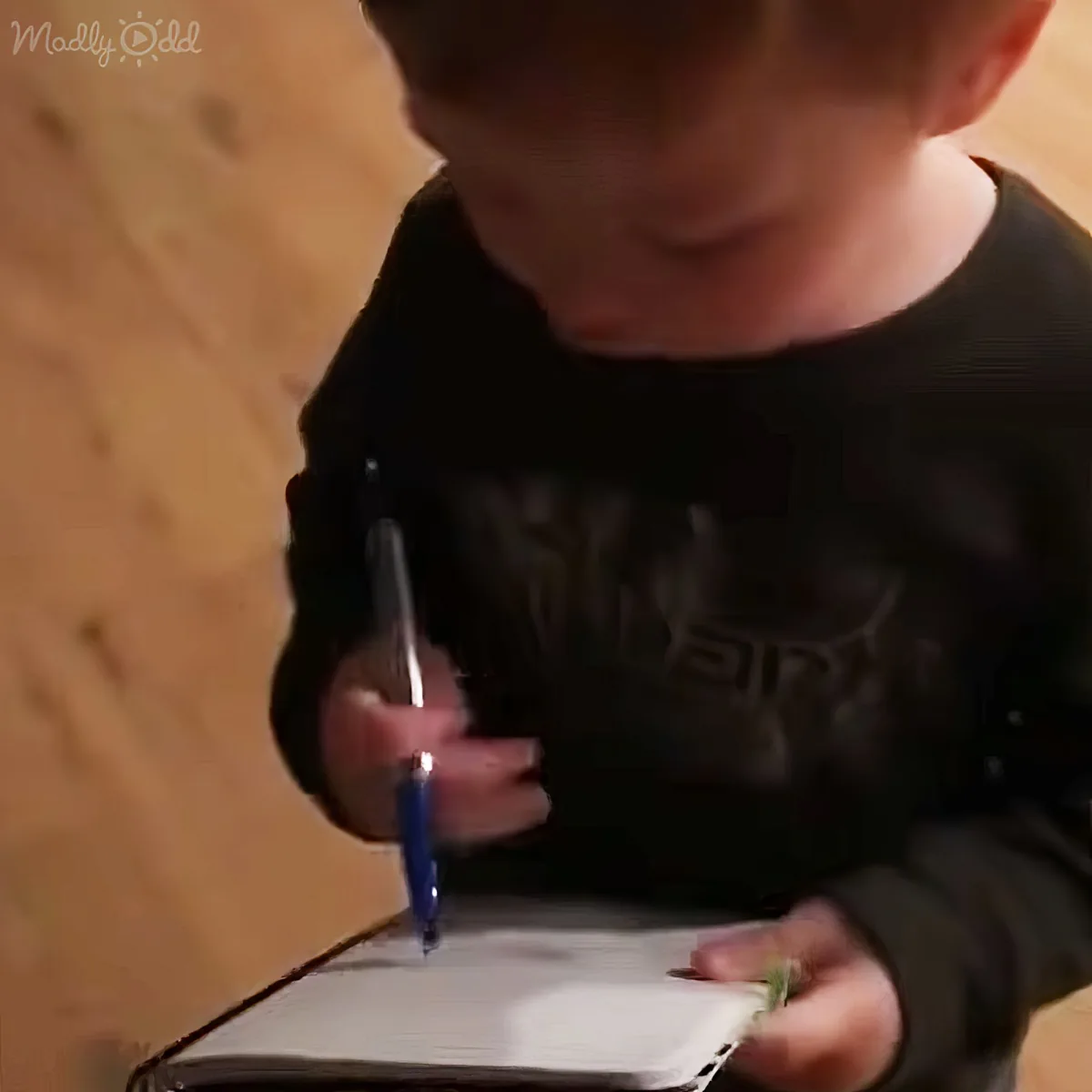 A young child in a black shirt is focused on writing in a notebook with a blue pen.