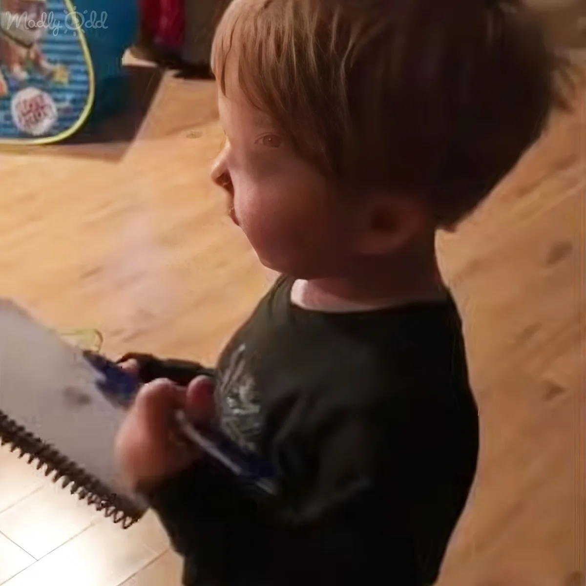 A young child with short blonde hair, wearing a dark shirt, is holding a toy guitar and looking to the side.