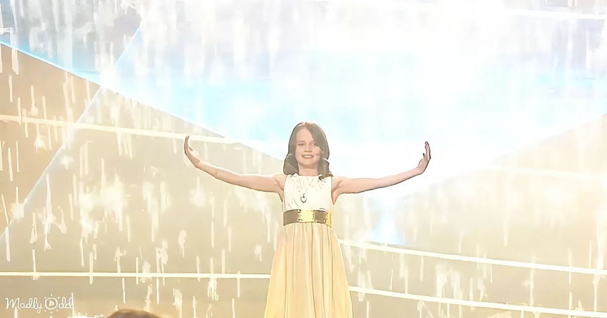 A person in a white and gold dress stands with arms outstretched, smiling, against a bright, glowing background.