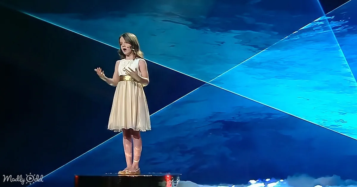A woman in a white dress stands on a stage with blue lighting, singing with her hands raised.