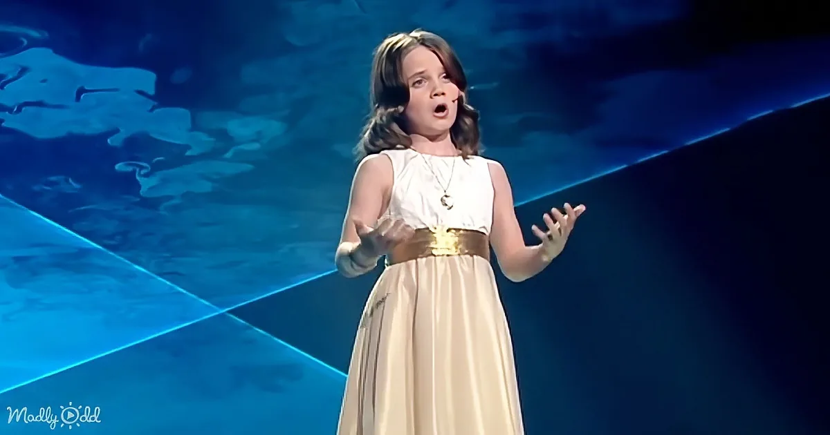 A young girl in a white dress with a gold belt sings on stage with a blue background.