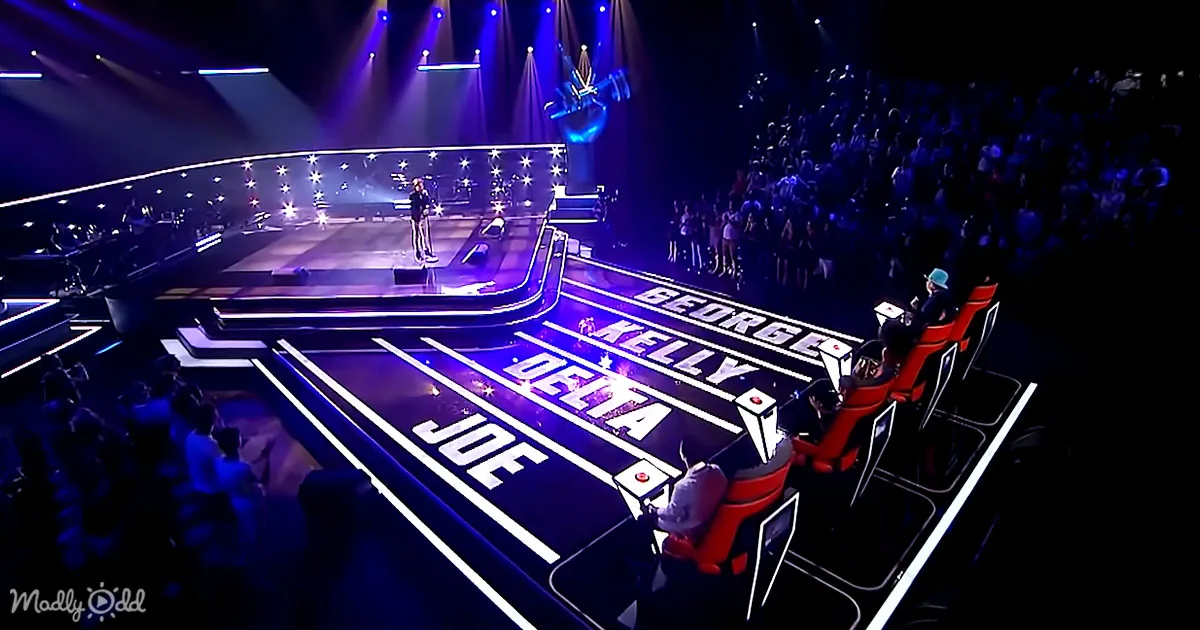 A contestant stands on a stage with judges' chairs labeled George, Kelly, Delta, and Joe, under vibrant stage lighting.