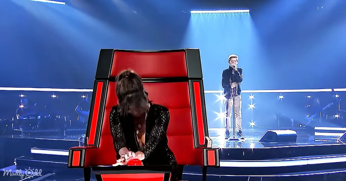 A person in a black outfit sits on a red chair, pressing a button, while another person stands on stage with a microphone.