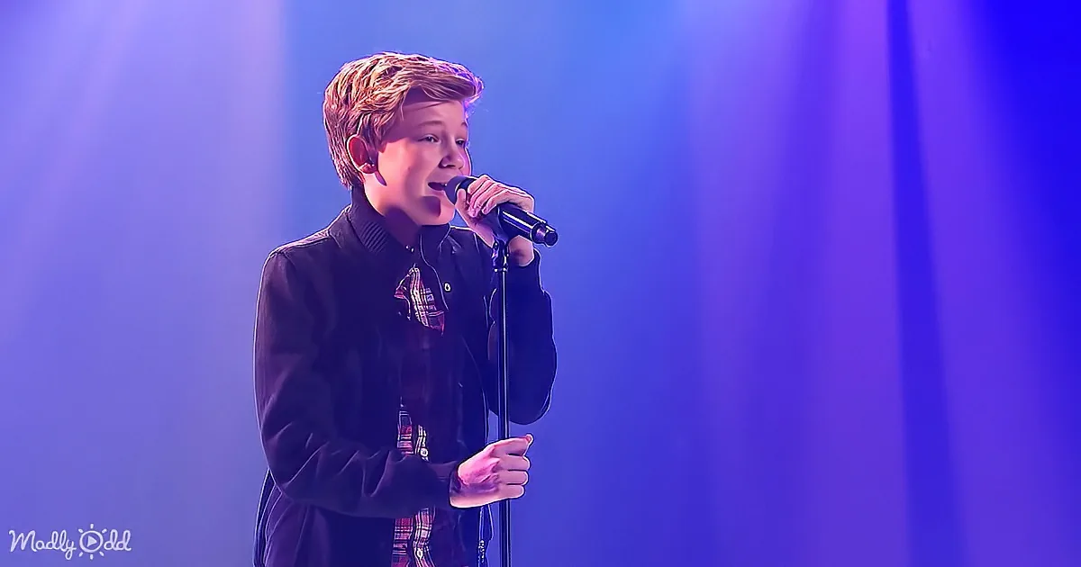 A young boy in a dark jacket sings into a microphone on a stage with blue lighting.