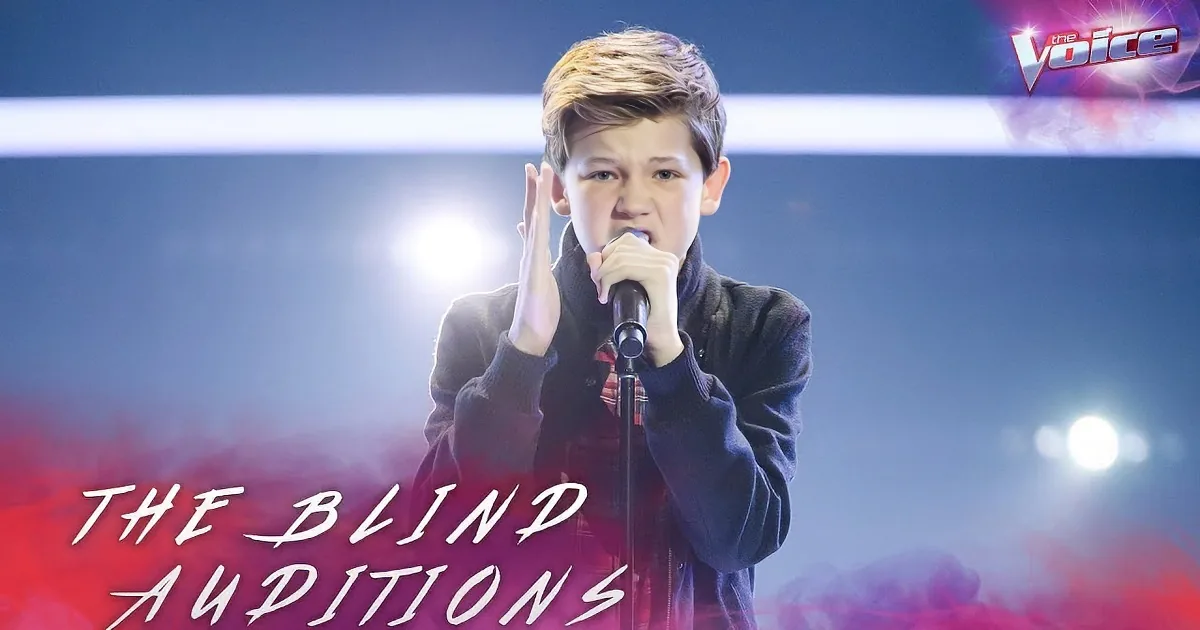 A young boy with short hair sings into a microphone, wearing a dark jacket and a red plaid shirt.