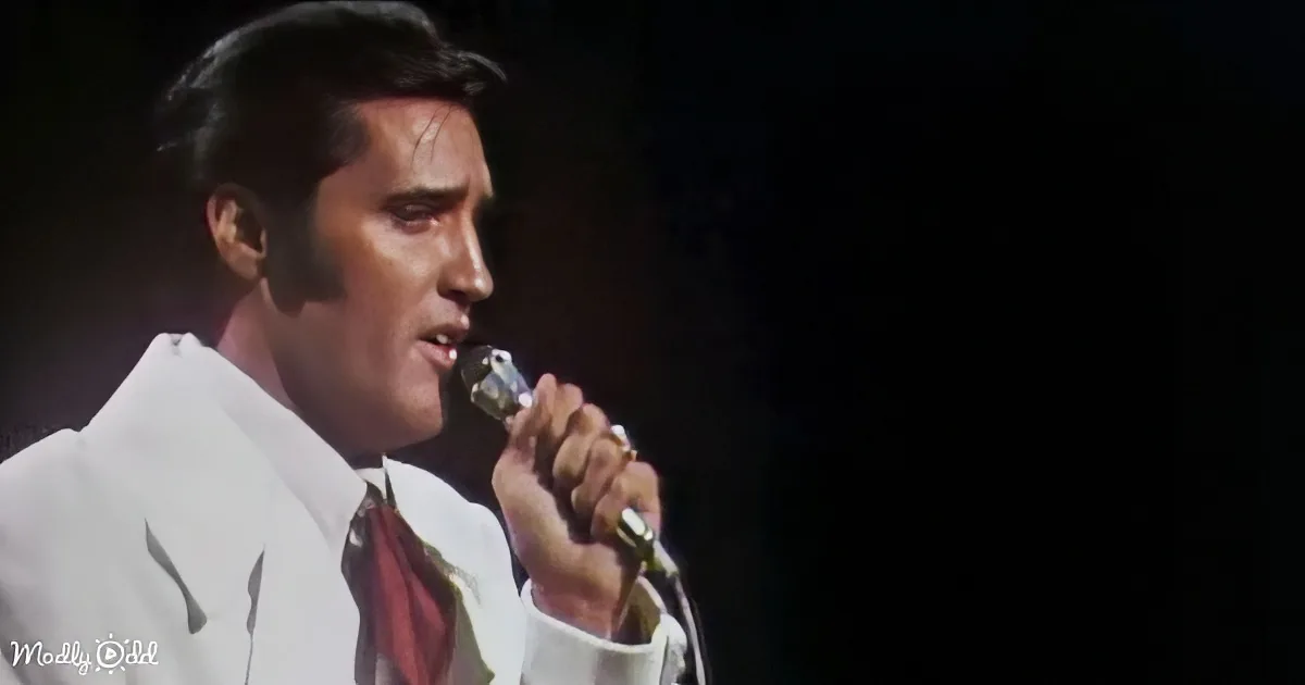Elvis Presley in a white suit and red tie sings into a microphone on stage.