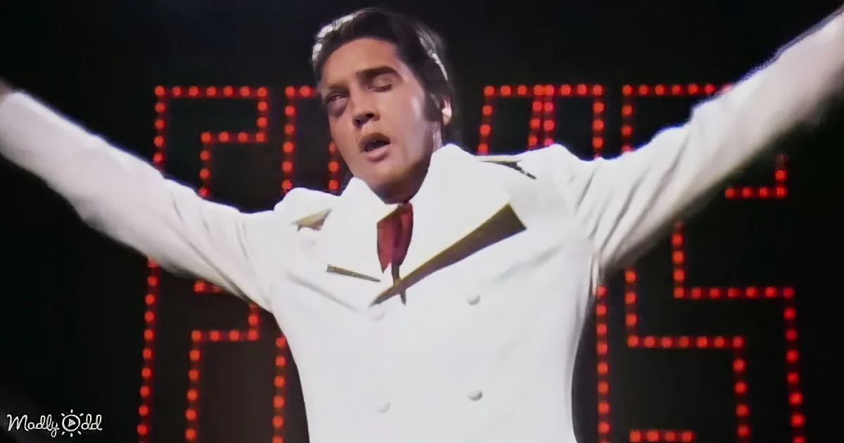 A man in a white suit with arms raised, standing in front of a red LED display.