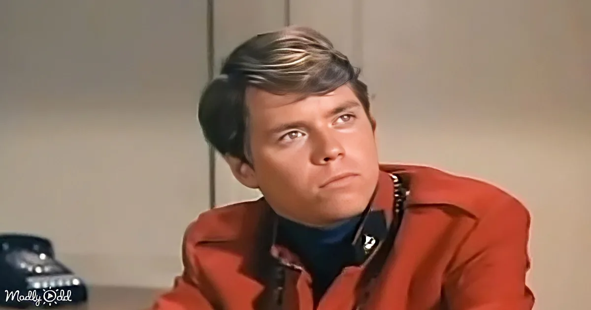A man in a red jacket looks up, wearing a headset, with a vintage telephone in the background.