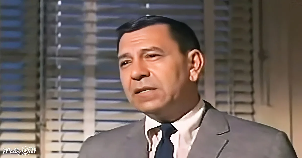 A man in a suit and tie stands in front of a window with blinds, looking to the side.