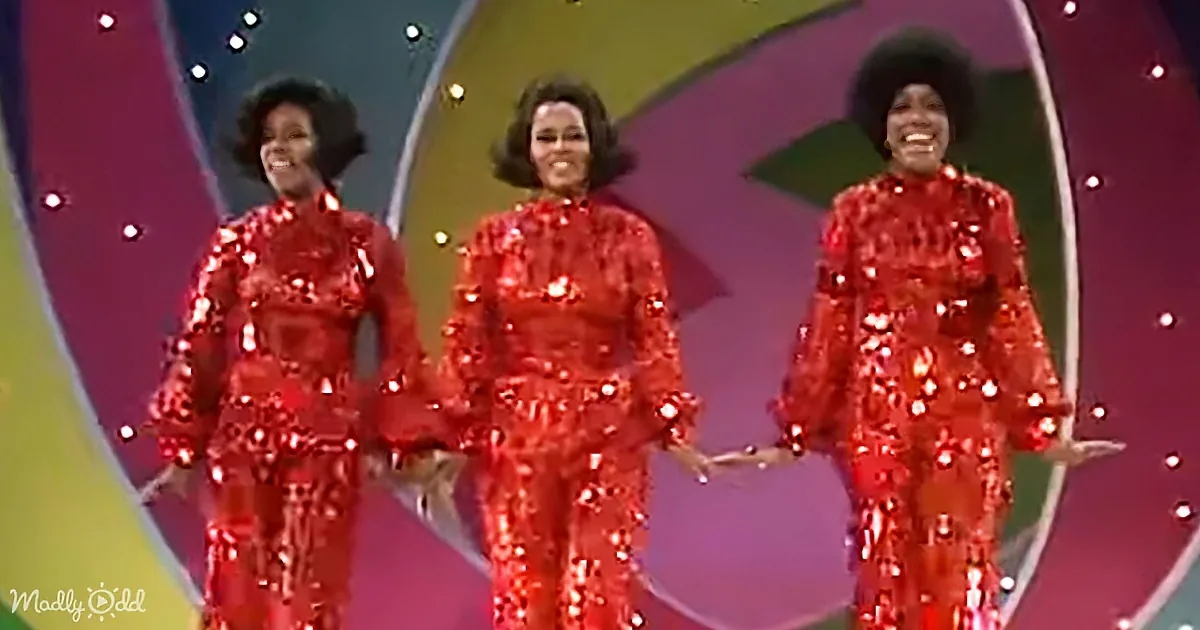 Three women in shiny red outfits perform on stage with colorful background.