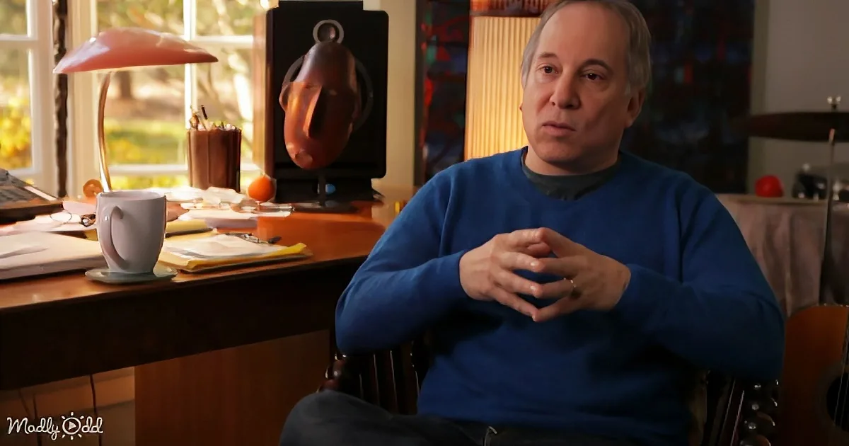 A man in a blue sweater sits in a chair, hands clasped, in a room with a desk, lamp, and guitar.