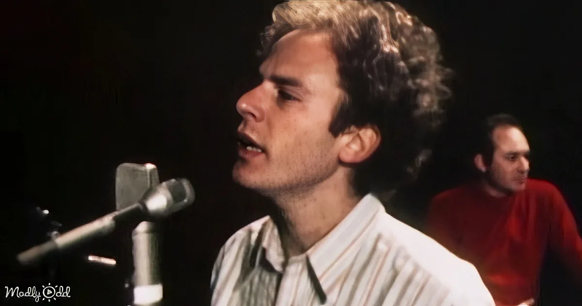 A man with curly hair sings into a microphone, wearing a striped shirt, with another person in a red shirt in the background.