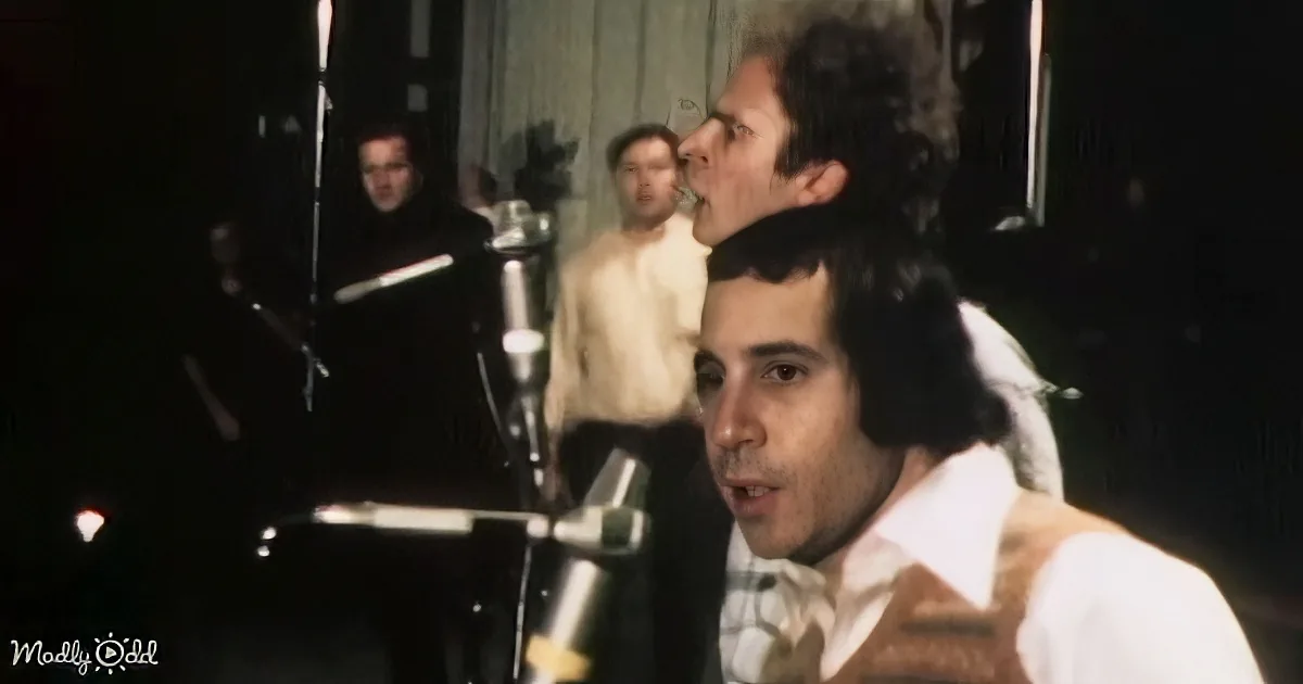 A man in a white shirt and brown vest sings into a microphone, with a band and drum set in the background.