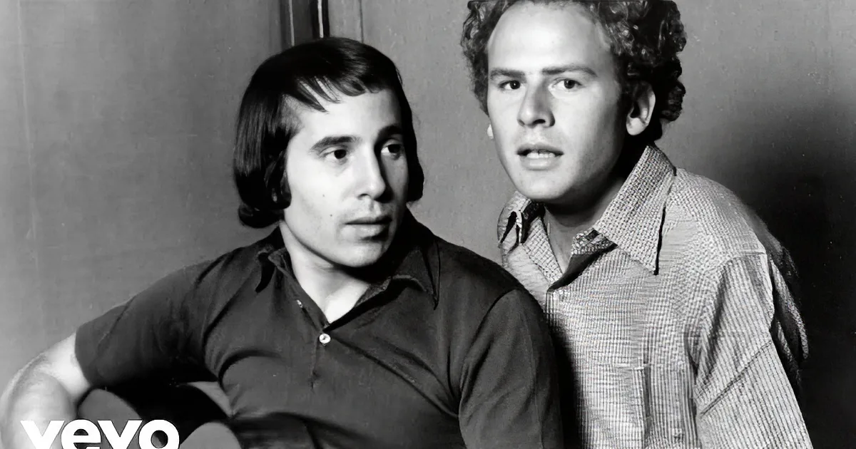 Two individuals in casual shirts, one with a guitar, posed together in a black-and-white photo.