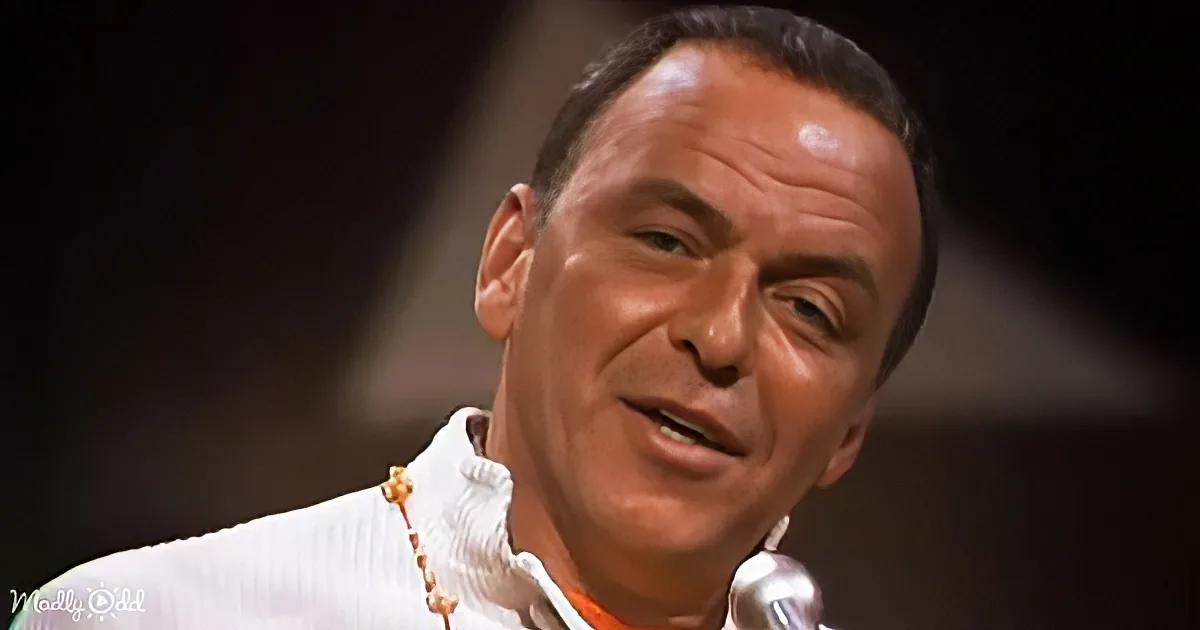 A man with short dark hair, wearing a white jacket and an orange shirt, is speaking with a serious expression.