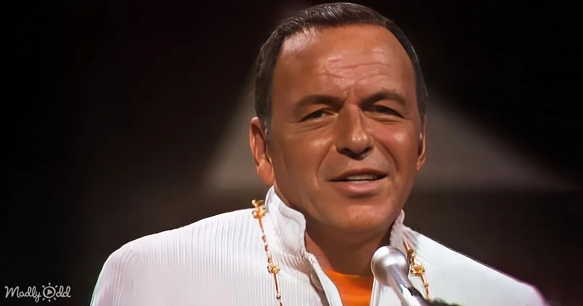 A man in a white shirt with a microphone, smiling, and wearing a necklace.