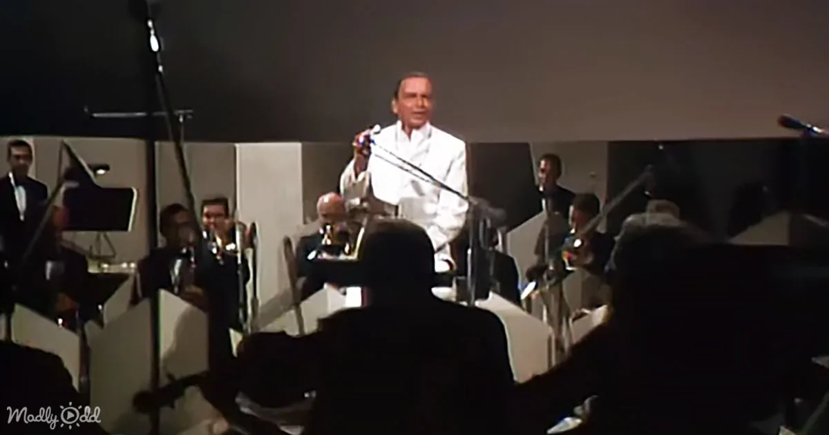 A man in white stands on stage with a microphone, surrounded by musicians in a dimly lit room.