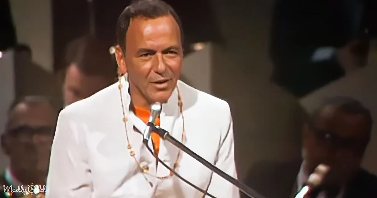 A man in a white suit and orange shirt speaks into a microphone, surrounded by blurred figures.
