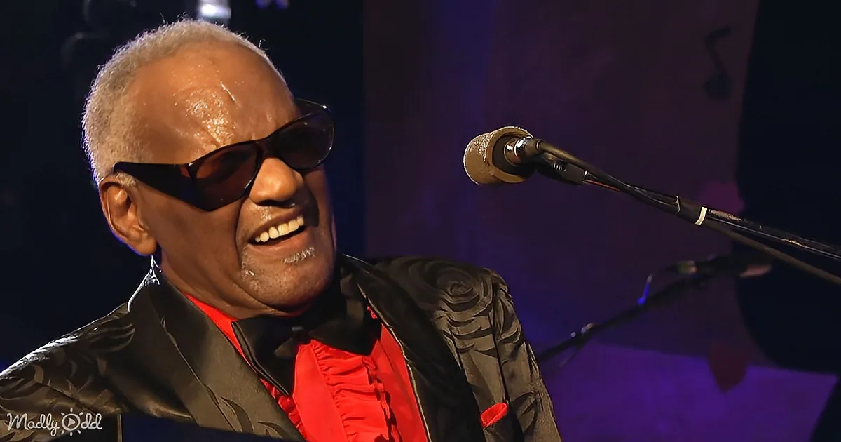 A smiling man in sunglasses and a red shirt performs on stage, wearing a patterned jacket.