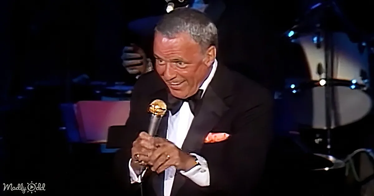 A man in a tuxedo holding a microphone, smiling, with a drum set in the background.