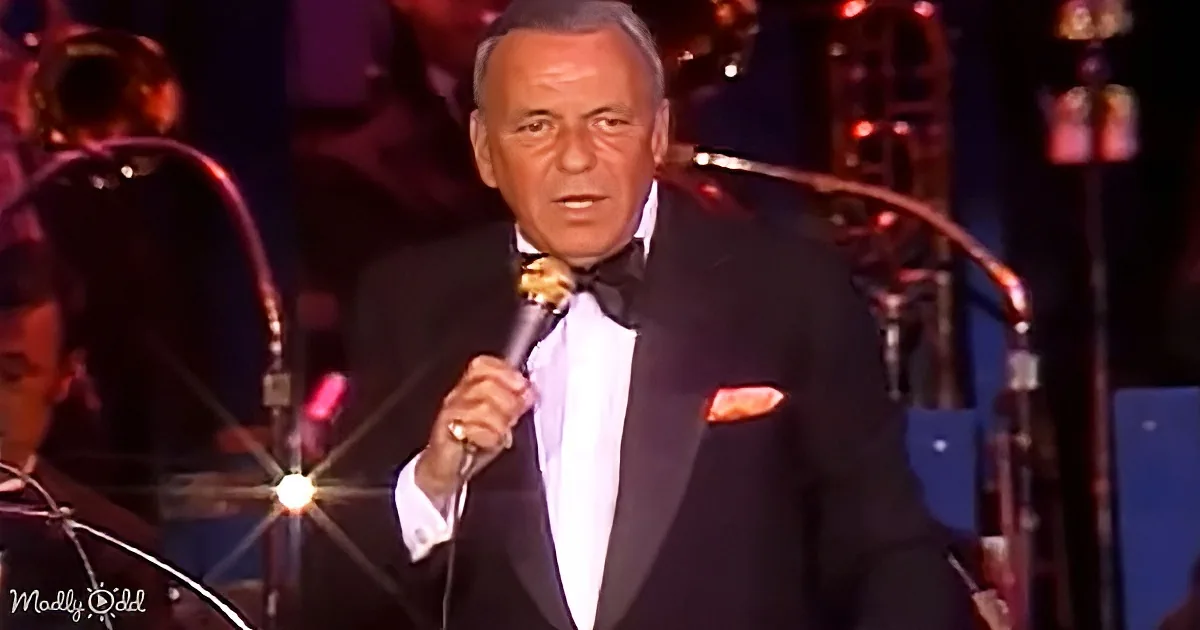 A man in a tuxedo holds a microphone, singing on stage with a band in the background.