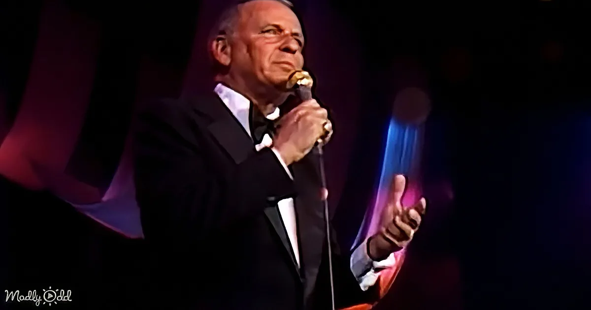 A man in a tuxedo holds a microphone, gesturing with his other hand on stage.