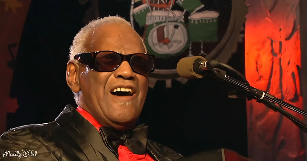 A man wearing sunglasses and a black suit sings into a microphone, with a colorful drum set in the background.