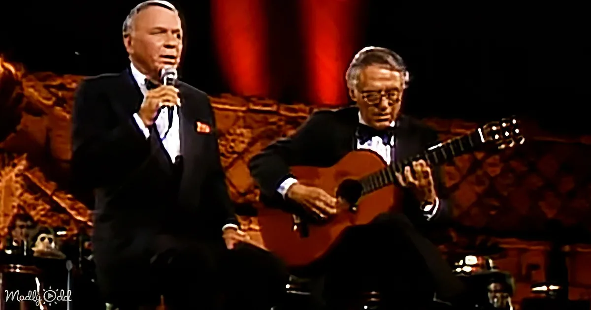 Frank Sinatra sings while playing a microphone, accompanied by a guitarist in a suit.