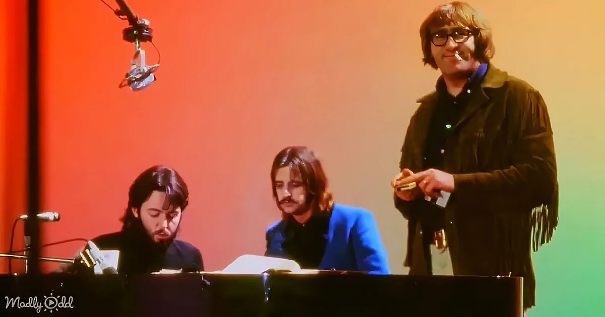 A man in a brown jacket stands holding a microphone, while two others sit at a keyboard, one with long hair and a mustache.
