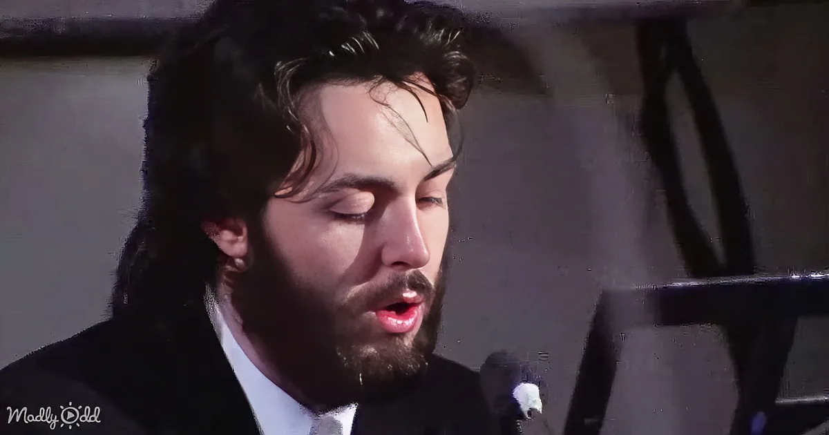 A man with long hair and beard sings into a microphone, eyes closed, wearing a suit.