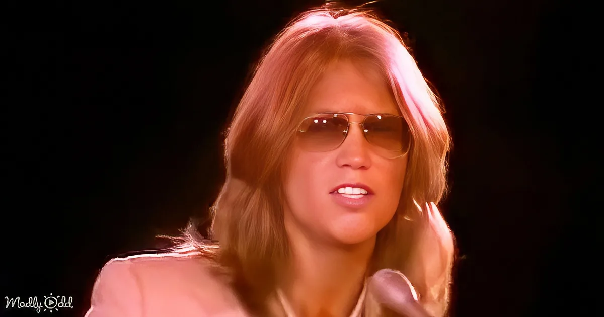 A person with long hair and sunglasses is seen in a close-up shot, wearing a light-colored outfit.