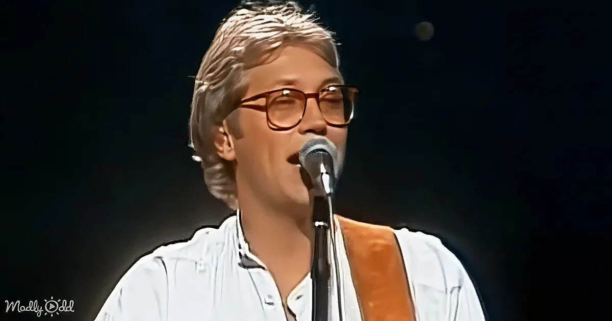 A person wearing glasses and a white shirt sings into a microphone on stage.