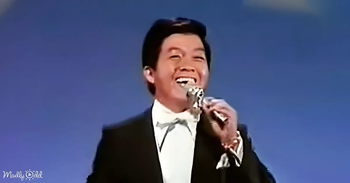 A man in a tuxedo smiles while holding a microphone, set against a blue background.