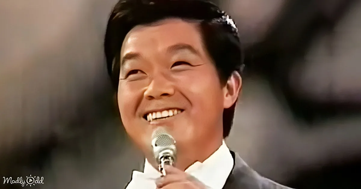 A smiling person in a suit holding a microphone, likely performing or speaking.