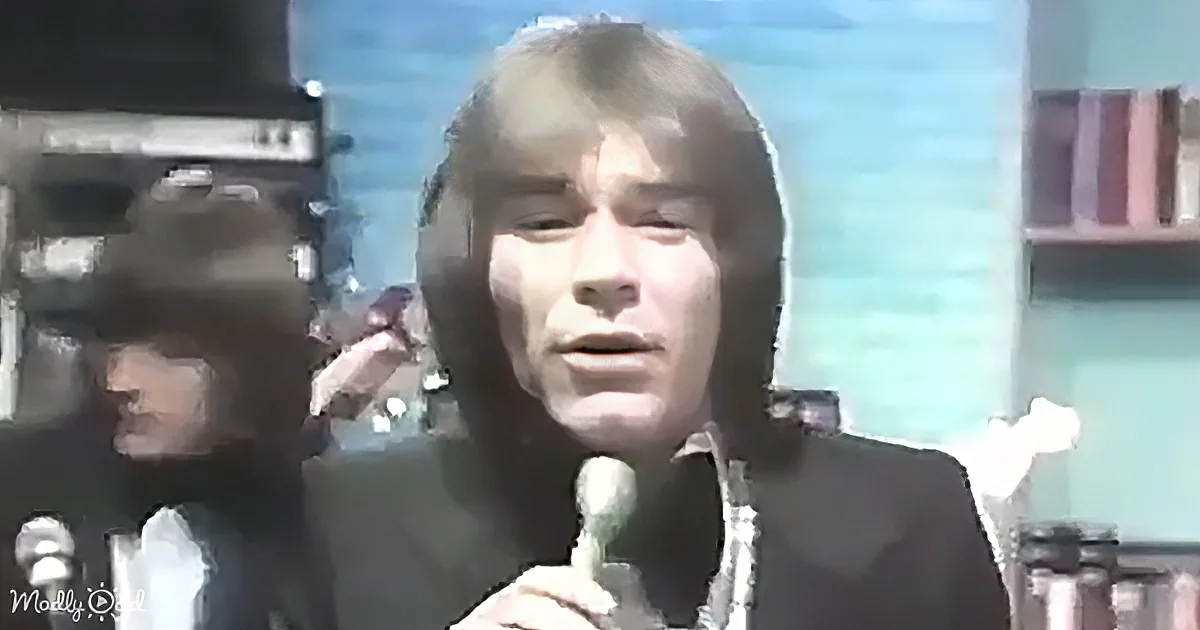 A person with long hair holds a microphone, possibly singing or speaking, in a setting with a blurred background.