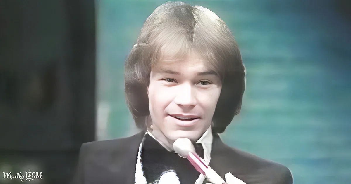 A person in a suit holding a microphone, smiling, with a blurred background.