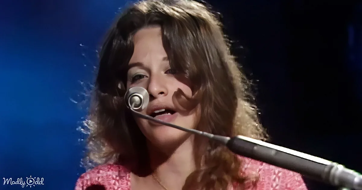 A woman with long, wavy hair sings into a microphone, wearing a pink patterned top, against a dark blue background.