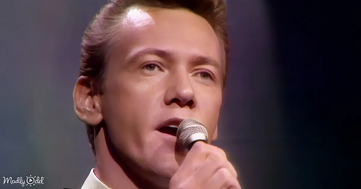 A man with short, styled hair singing into a microphone, wearing a white shirt.
