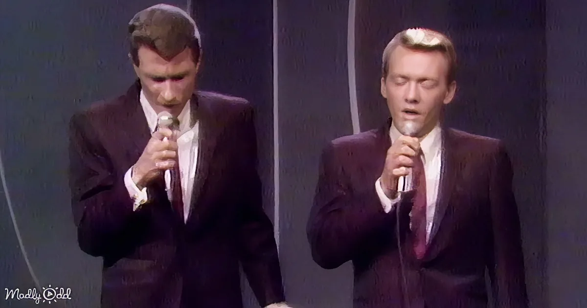 Two men in suits sing into microphones on stage, one with a receding hairline.